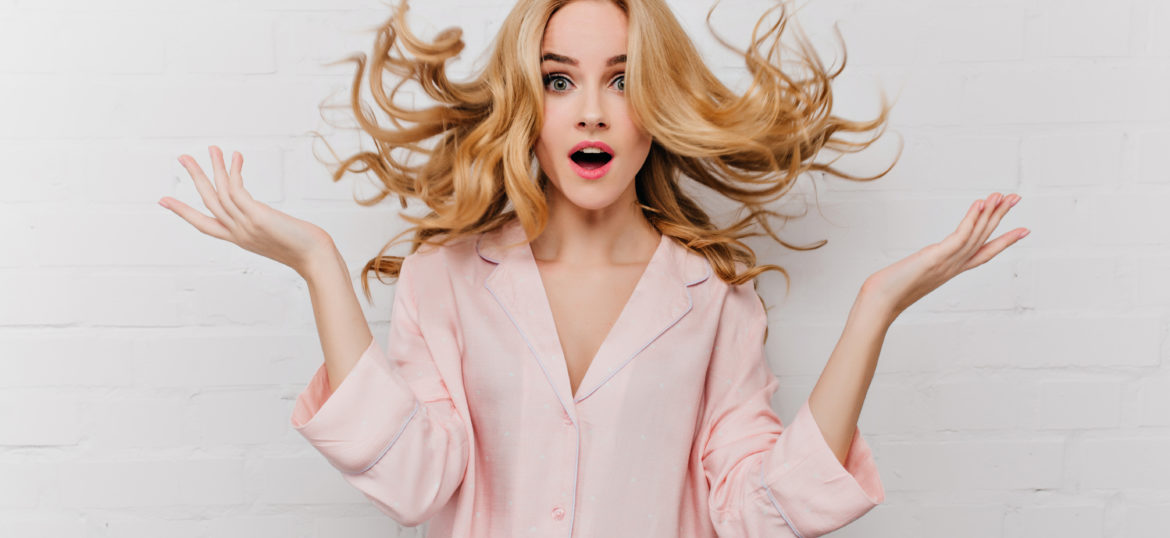 Ecstatic blue-eyed woman with long blonde hair posing in front of white bricked wall. Indoor shot of surprised girl in beautiful pink pyjamas..